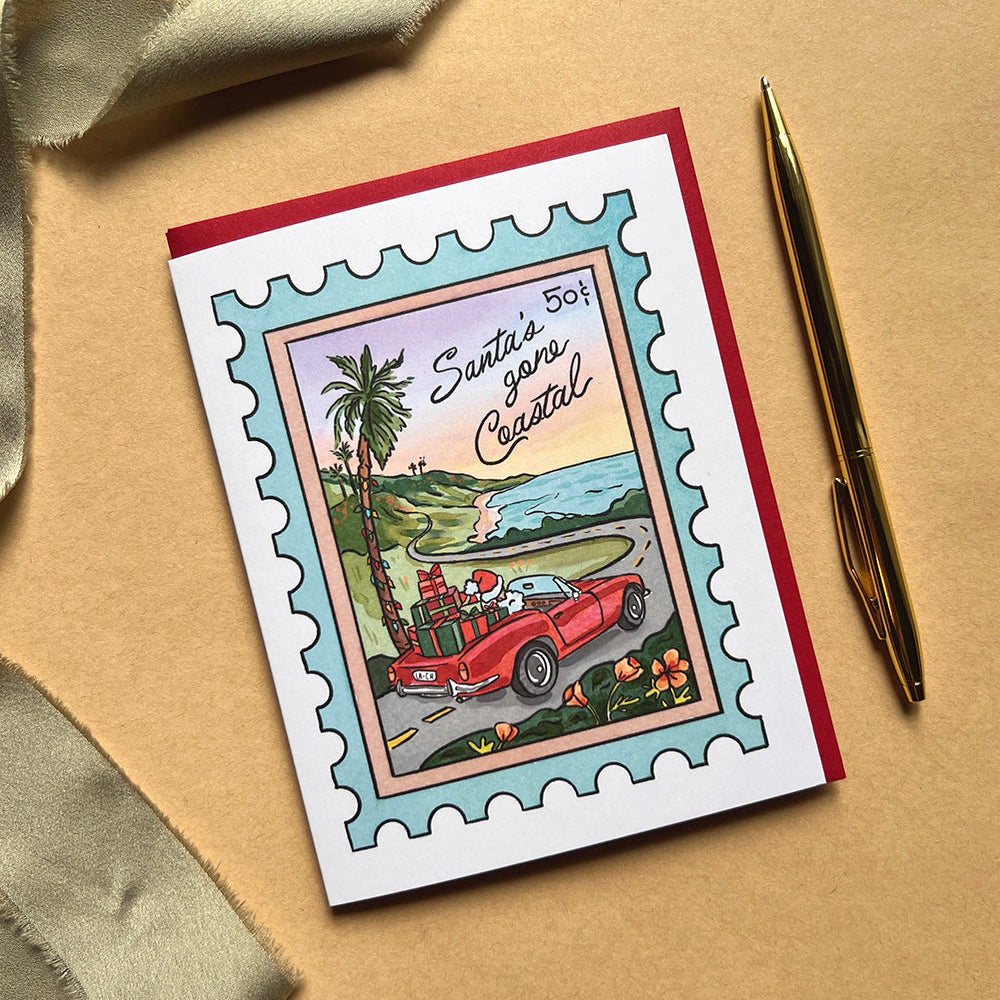 Greeting card with a tropical design featuring a red car and palm trees, placed on a beige surface with a gold pen and ribbon.
