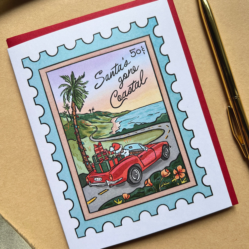 Greeting card with Santa driving a red car and palm trees, on a beige surface with a pen.