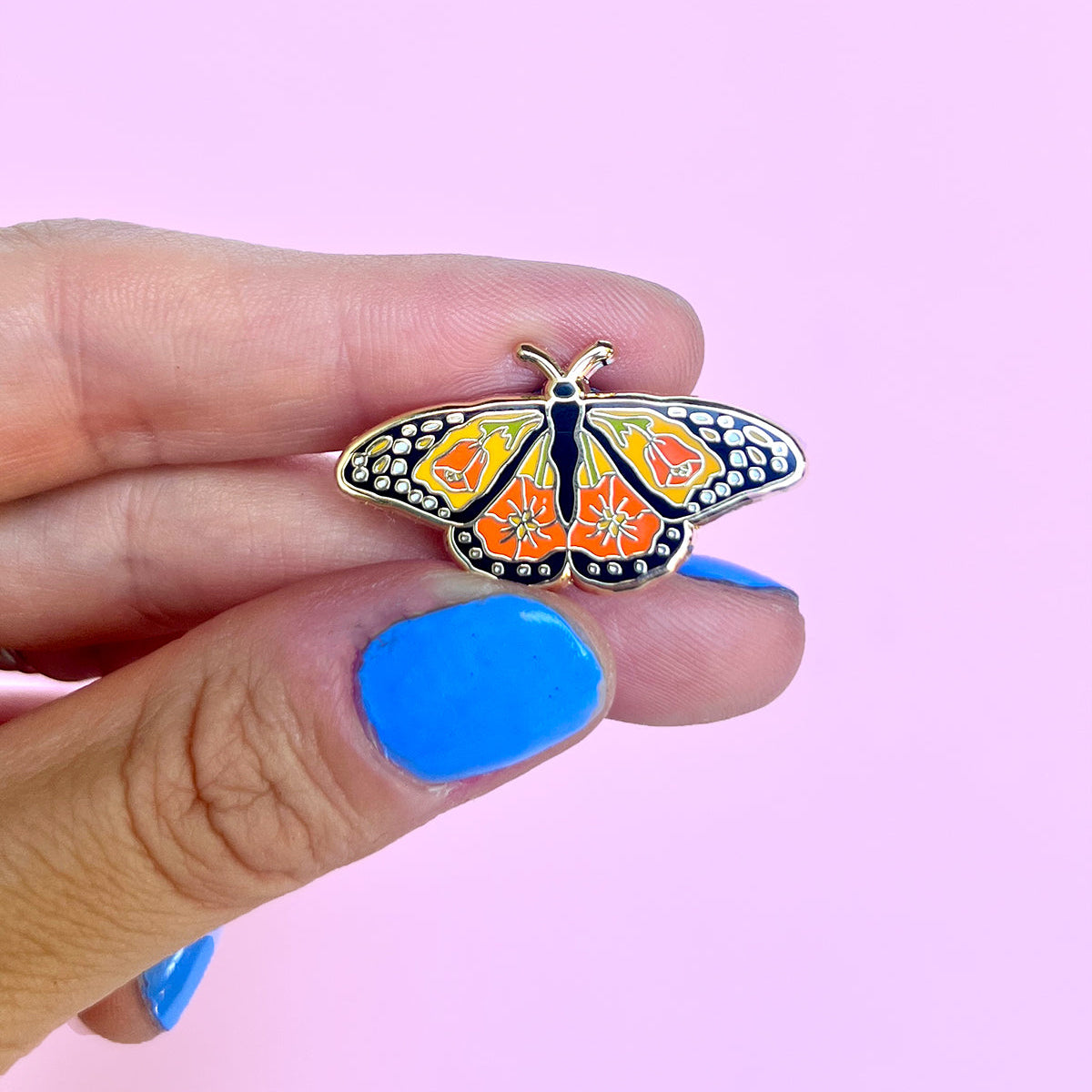 Monarch butterfly-shaped pin held by a hand against a pink background