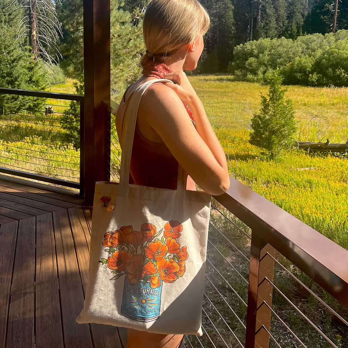 Person holding a tote bag with a poppy design, standing on a wooden deck with a scenic background of trees and fields.
