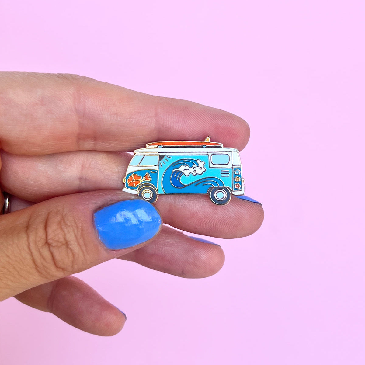 Hand holding a colorful surf van enamel pin against a pink background