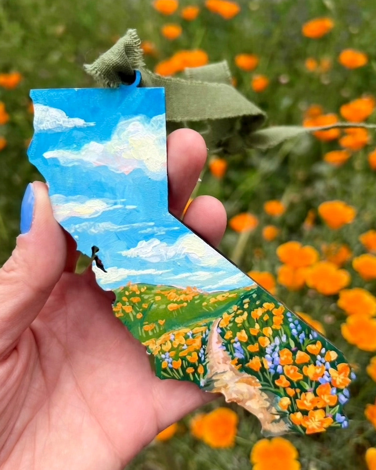 Hand holding a painted wooden state with a scenic landscape, surrounded by orange flowers.